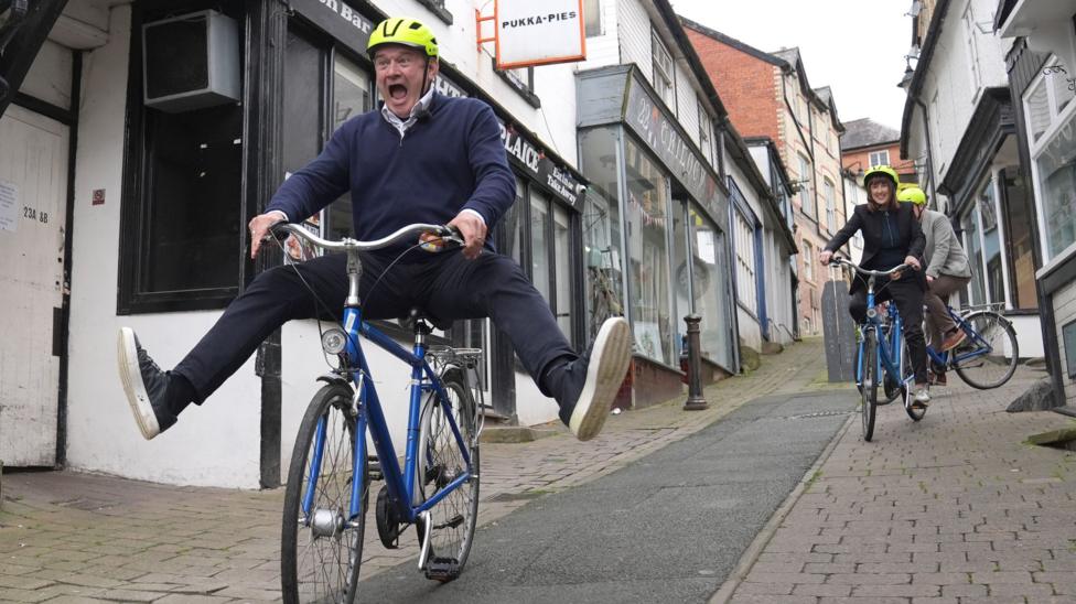 Ed Davey riding a bicycle with his legs open as it goes down a hill. The bicycle is blue. Jane Dodds is riding a bike in similar colours and is behind him. They are wearing yellow helmets.