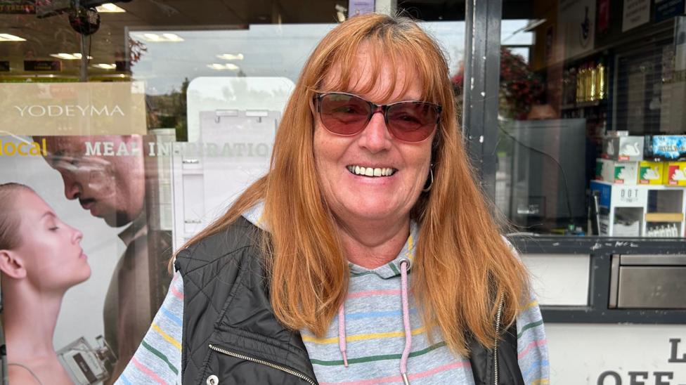 Sue Willis stands in front of a shop window. She is smiling and has long auburn hair with a full fringe. She is wearing black sunglasses with red lenses. She is wearing a grey hoody with thin, rainbow coloured stripes and over the top of it is a black body warmer.