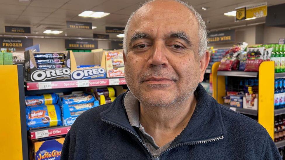 Shailesh Parekh stands in the convenience shop attacked to his petrol station. Behind him are rows of shelves holding biscuits, crisps and drinks. He is an older man with grey hair and darker eyebrows. He is wearing a dark, navy zip-up fleece and underneath can be seen a grey polo shirt.