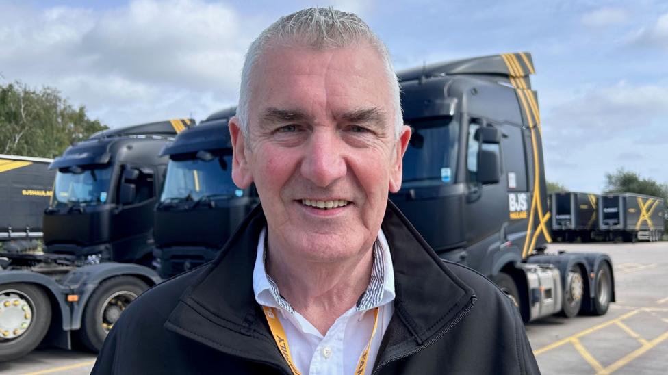 Davide McWilliams is an older man with short grey hair, he is looking at the camera and smiling. He is wearing a black zip-up jacket, white shirt and orange lanyard. Behind him are several black BJS Haulage trucks.