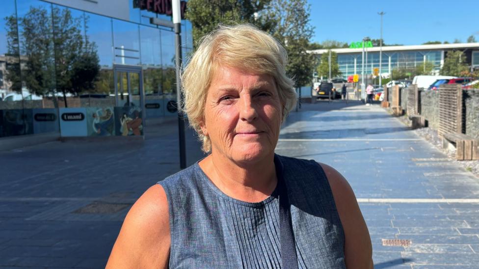 A woman with blonde short hair. She is wearing a blue sleeveless top with a black strap where a bag would be. She is stood on a black brick path with benches against the wall. In the background is a big carpark with a building that says 'ASDA' at the top.