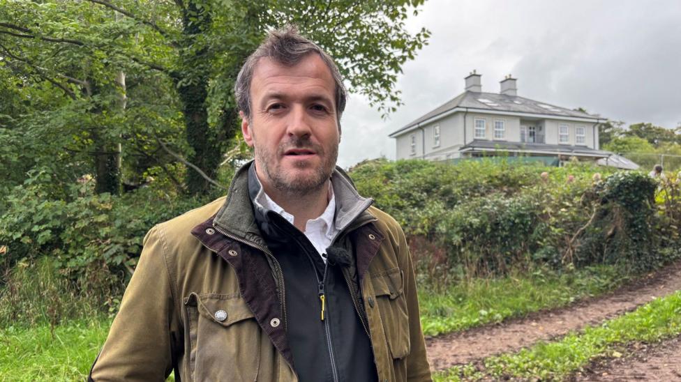 Emyr Davies standing on a small path in a green waxed jacket over a dark zipped jumper and white shirt. He has short dark hair and stubble. There is a large house in the background behind fencing.