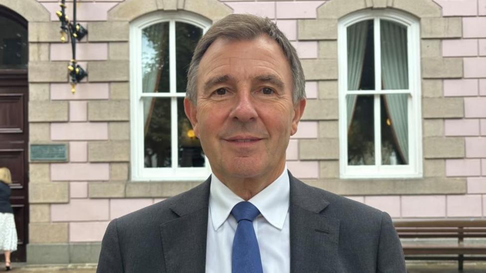 A person with short brown-grey hair stands faces the camera outside a stone-fronted building with pale pink walls and large sash windows. The person is wearing a dark grey suit jacket, white shirt and blue tie. The photograph is taken outdoors in daylight and is framed as a head-and-shoulders portrait.