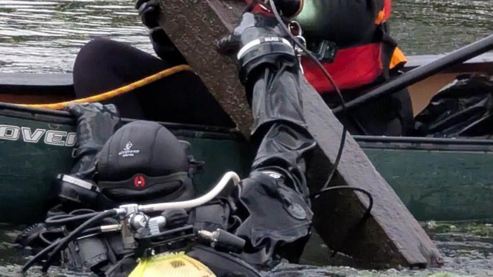 A diver in full diving gear which is all black and with an oxygen tank on their back emerges from the water with a large piece of wood. They are holding onto a green canoe, handing the wood to another person wearing red and black who is in the canoe. We can just see that person's lower body, as they receive the large plank of wood from the diver.