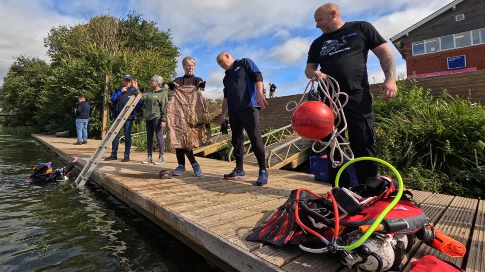 Six adults stand on a wooden jetty overlooking the river, with diving equipment piled up to the side. There's a diver emerging from the water, next to a ladder leading out of the river.