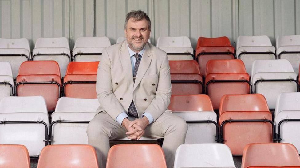 A man in a beige suit sits in an empty football stand, among red and white seats