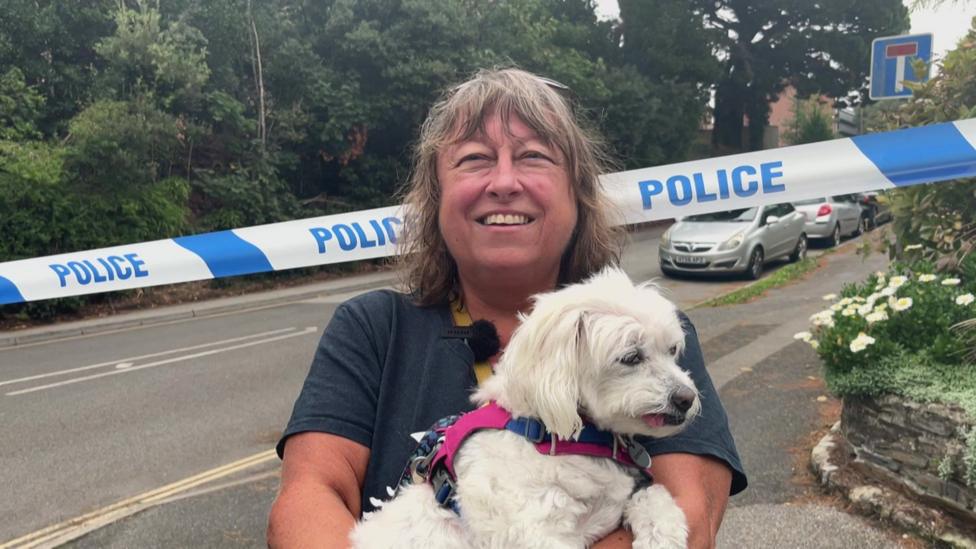 A woman holding a small white dog. There is police tape behind them and a street with parked cars.