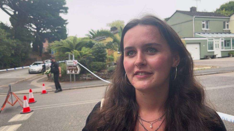 A woman with short brown hair. She has hoop earrings and a nose piercing and is wearing a black jacket. Behind her is police tape and a police officer. 