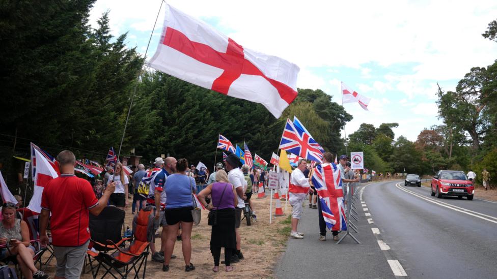 Protesters with St George and Union Flags stand beside a road.