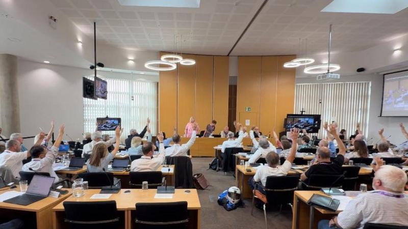 A meeting room at West Suffolk Council's HQ. There are rows of tables with people sitting at them, facing away from the camera. They are all raising their arms in a vote. At the far end of the room, there are at least five people at a desk, facing into towards the camera.