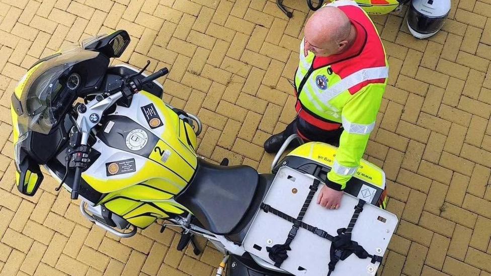 An aerial picture of a man standing next to a blood bike, which is covered in yellow hi-vis decals and equipped with medical supplies. The man is bald, wearing a yellow hi-vis and red vest, and leaning on the back of the motorbike.