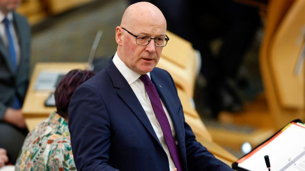 John Swinney, who is bald with glasses, wearing a business suit in a debating chamber