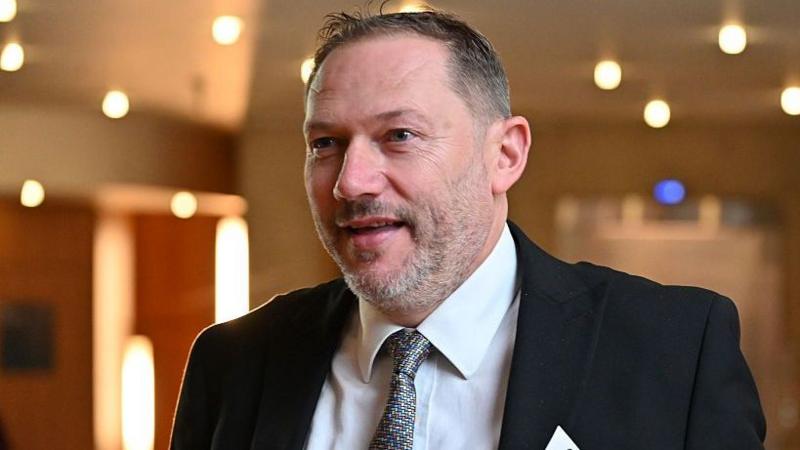 MP Douglas Lumsden, a man with a grey/black beard and short dark hair in a black suit with a grey shirt and patterned tie