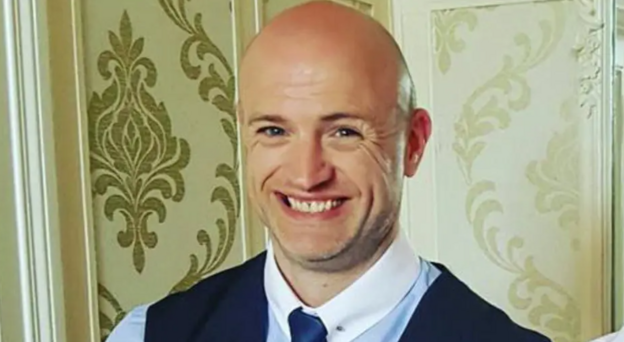 Shows a man with bald head smiling. He is wearing a blue shirt with shite collar and navy waistcoat. 