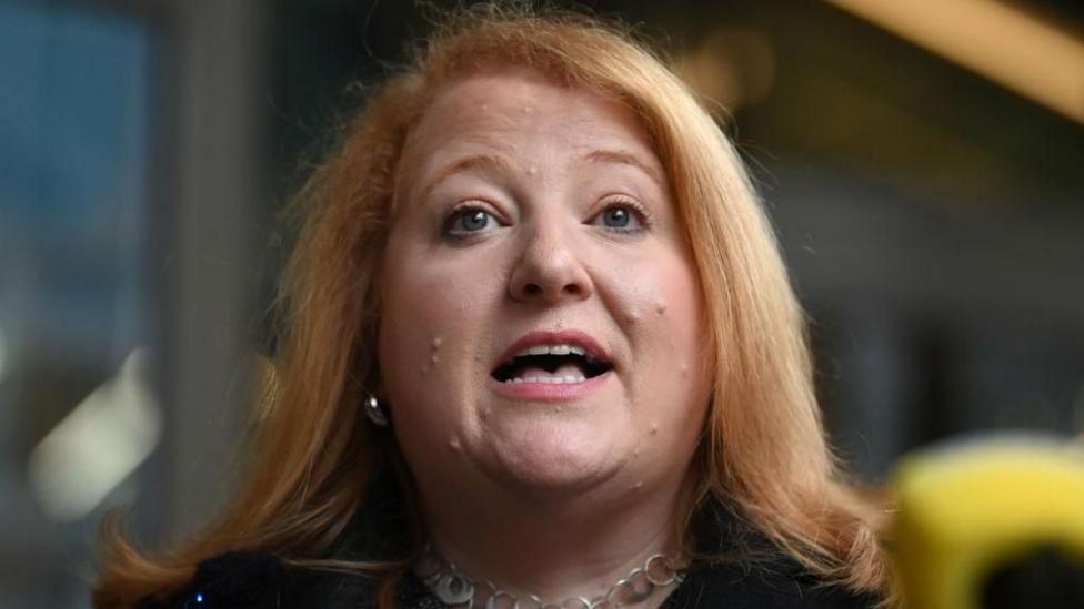 A red-haired woman with green eyes wears a black jacket and silver necklace. She is talking into a yellow microphone. She wears small silver earrings.