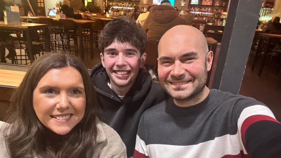 A selfie or Riley, with his mum and dad on either side of him. They all smile at the camera. Chris is bald with short stubborn, Kate has long dark hair. Riley has short dark hair and wears a black hoodie.