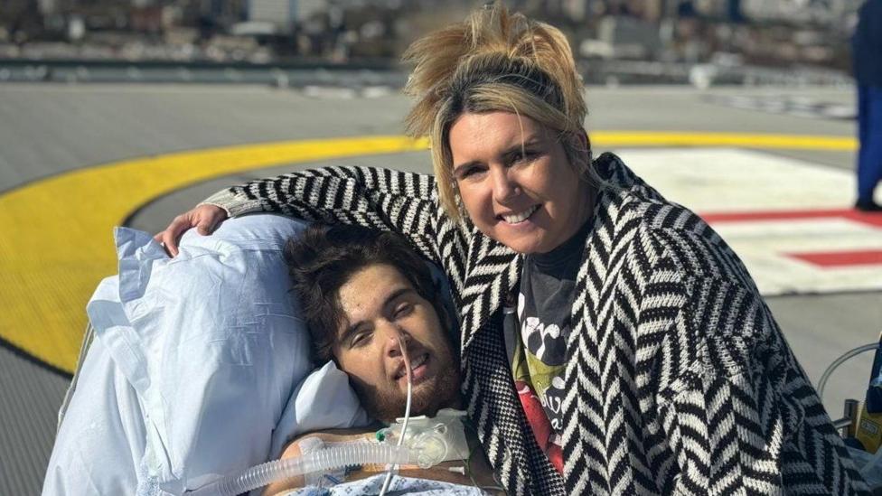 Riley in a hospital bed, with a tube connected to his nose. He has short dark hair. His mother has her arm around the bed, and has blonde hair which is tied back. She wears a patterned black and white cardigan. A square of concrete can be seen behind them.
