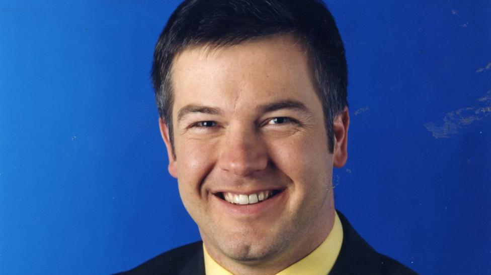 A young Derek Brockway has dark hair and is pictures against a blue screen and dark suit and yellow shirt in1997