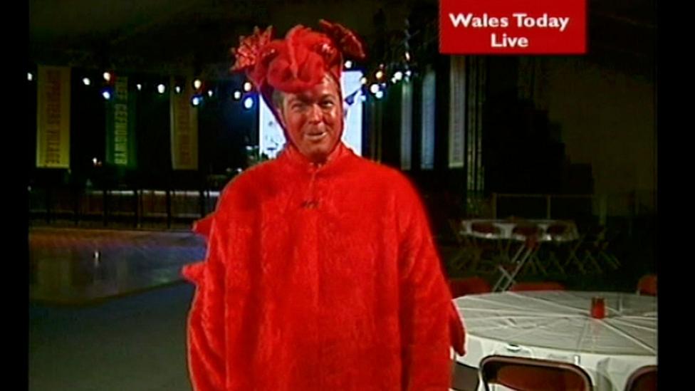 Derek wears a full red dragon fancy dress outfit and has a painted red face