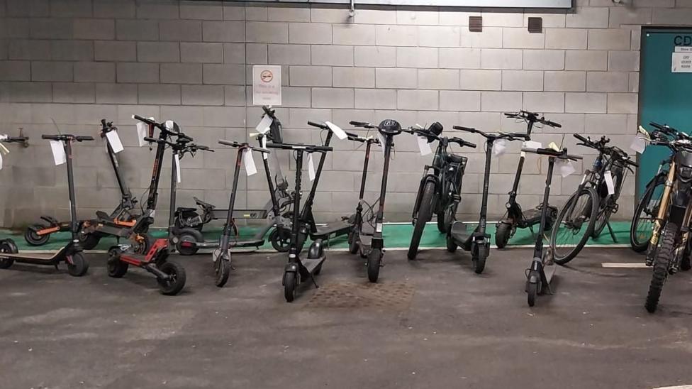 E-scooters tagged sitting on tarmac. Behind them is a grey brick wall with a blue door to the right