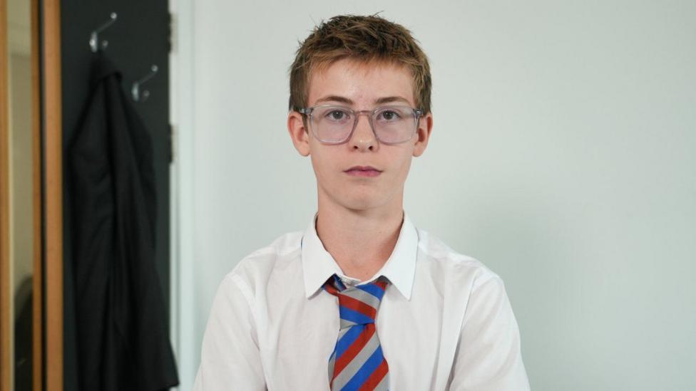 Harrison is wearing a white school shirt and striped tie. He is wearing glasses.