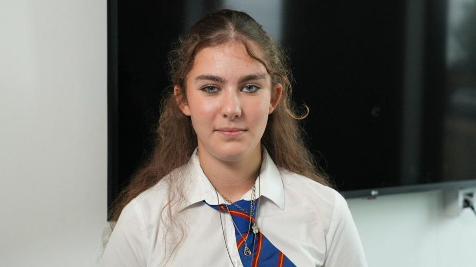 Anabella is wearing a white school shirt and blue tie, she has long brown hair.