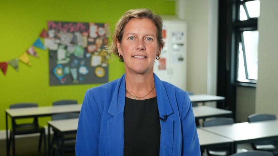 Henriette is wearing a black dress and blue blazer, she is stood in a classroom.