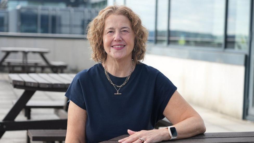 Mary-Ann is wearing a navy dress and has short, curly hair. She is sat at an outdoors bench.