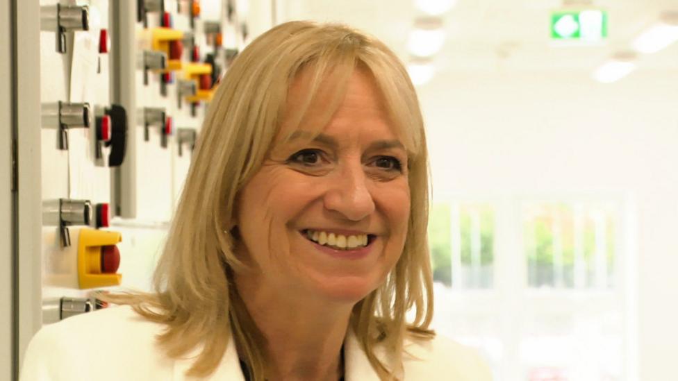 A blonde woman in a white jacket smiles at someone off-camera, behind her are distant windows in a white corridor. Over her shoulder are a number of red emergency stop buttons.