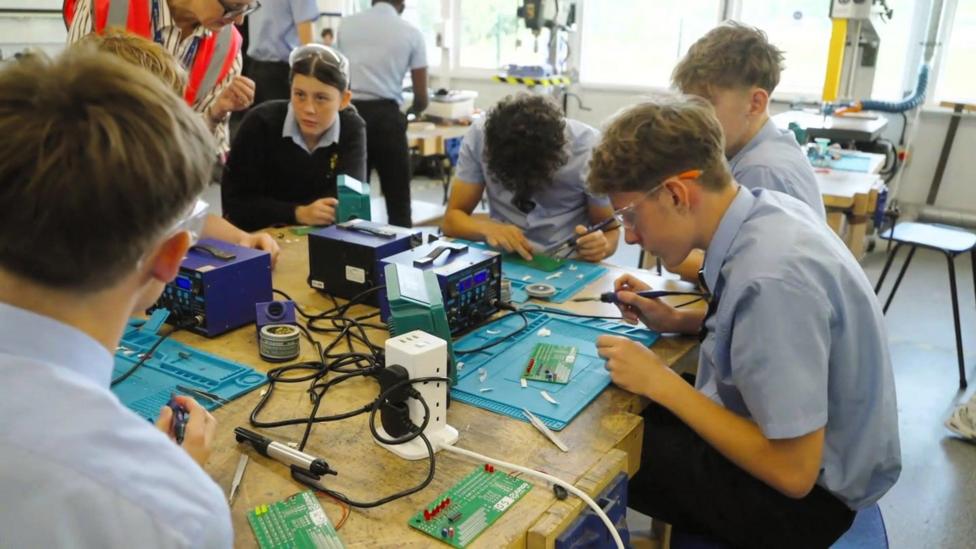 A group of students sit around a wooden table soldering circuit boards during a lesson