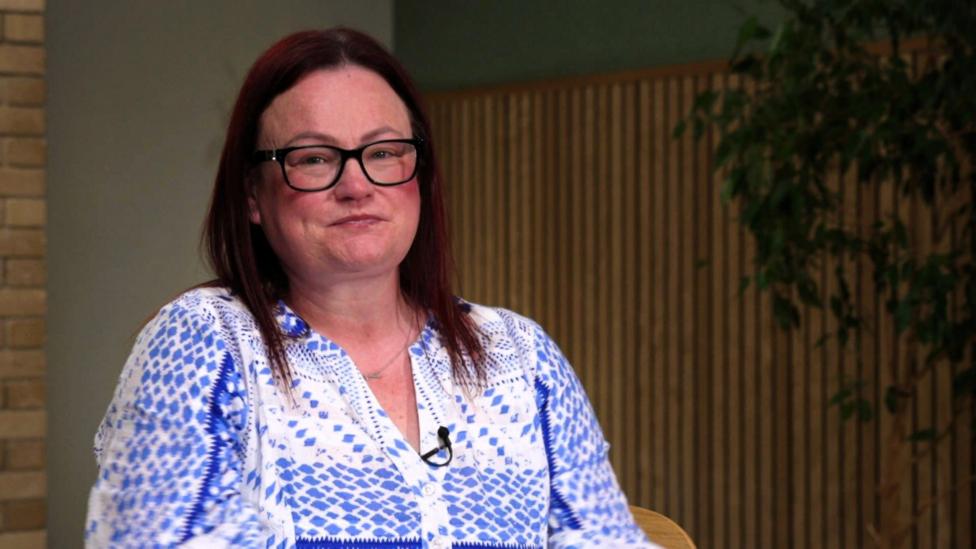 A dark-haired woman in glasses and a blue top smiles at the camera. The office backdrop includes wood panelling and the green leaves of an indoor plant.