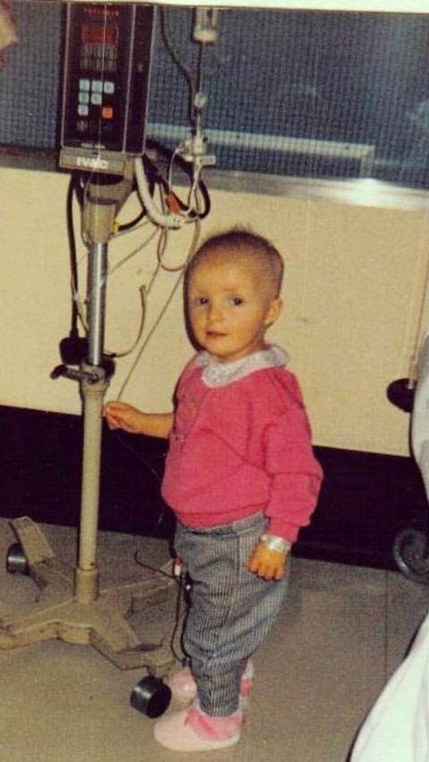 Nicole Anderson as a very young child. She is standing in a hospital ward, aged about three or four, wearing a pink jumper and blue toursers and pink shoes. She is standing next to a machine with a drip in it, and is presumably being treated that way.