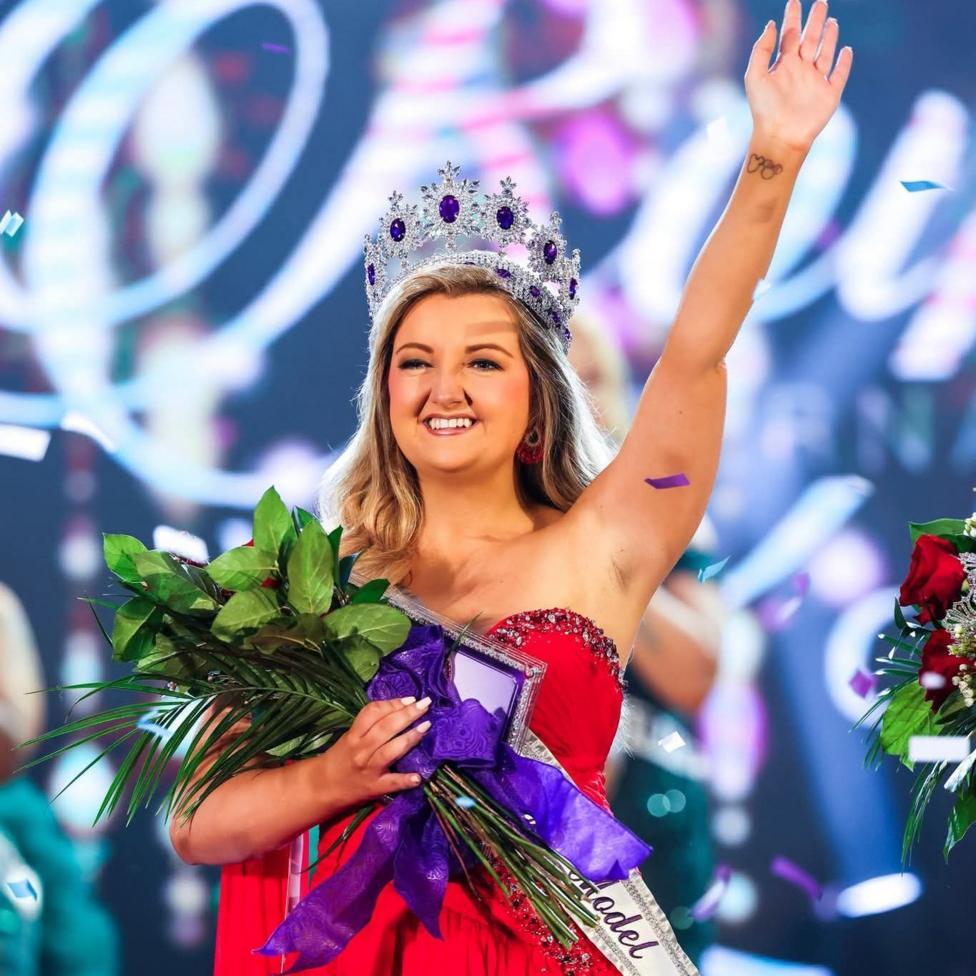 Nicole Anderson stands on a brightly lit stage wearing a jeweled tiara, a red evening gown, and a sash. The contestant holds a bouquet of green foliage tied with a large purple ribbon in one arm while raising the other arm overhead. Colourful confetti falls around the stage.
