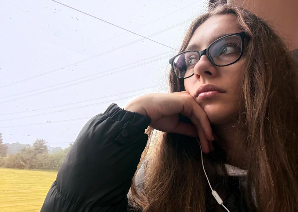 A young woman with long brown hair and dark-rimmed glasses stares out of a train window as it moves along. She is wearing a dark jacket and white earphones are visible.