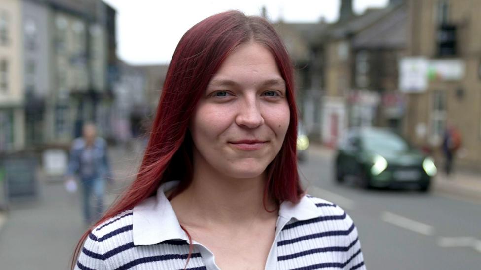 A young woman with long hair and a stripy top stands on a pavement in Pennistone