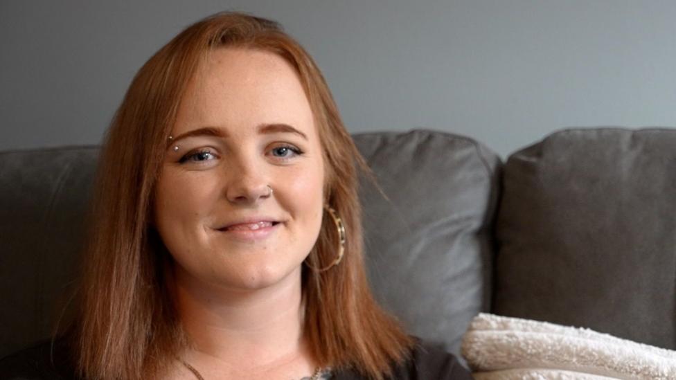 A woman with shoulder-length ginger hair and a black top smiles as she sits on a sofa.