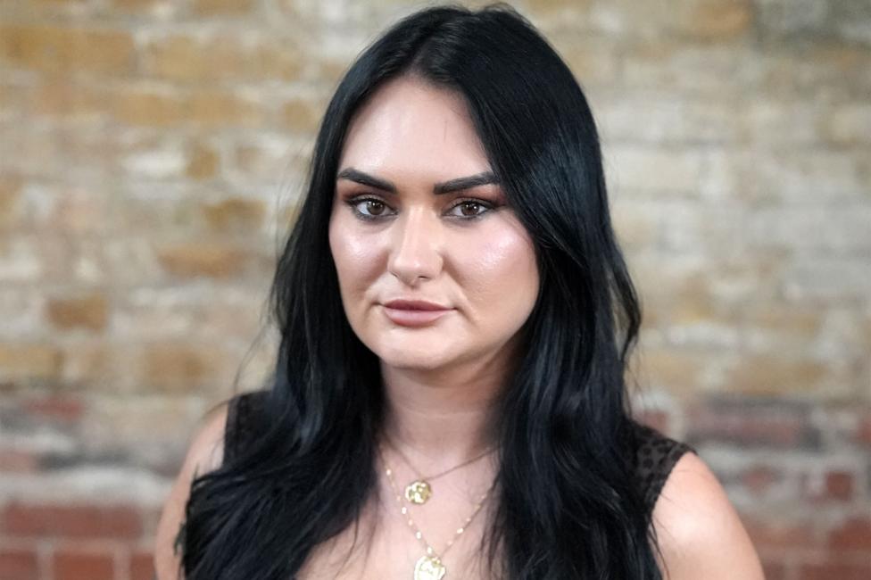 Nikita has long dark hair, wears a sleeveless top and two gold necklaces, and is standing against a textured brick wall.