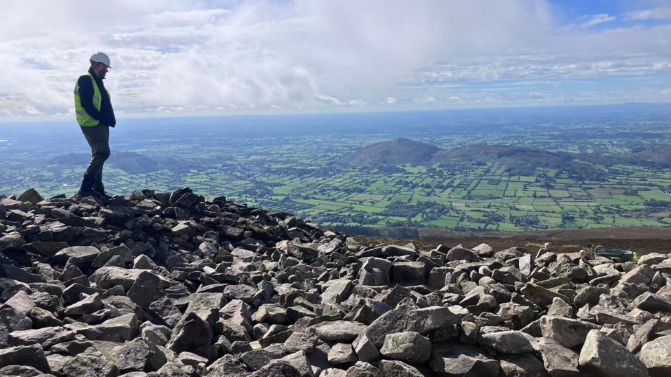 A man stands a top large stones and boulders overlooking fields, moutains. Above is blue sky and fluffy clouds