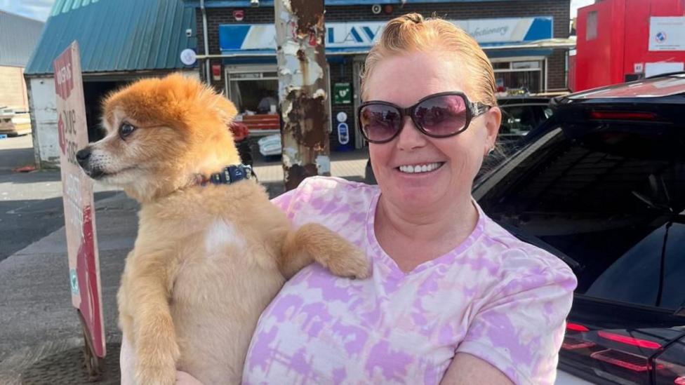 Irene is smiling at the camera and is wearing large sunglasses and a purple tie-dye t-shirt. She is holding her dog which is a Pomeranian type and a light brown/ginger colour.