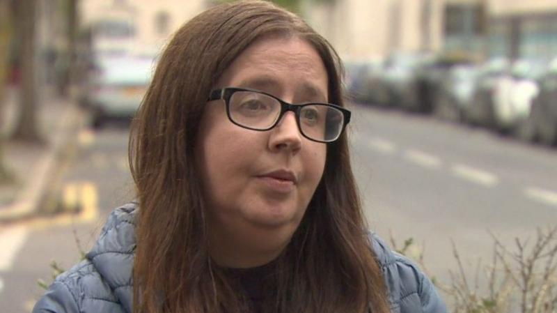 Michelle Kelly has long brown hair parted in the middle and black-rimmed glasses. She is standing in a city street wearing a navy coat and dark top
