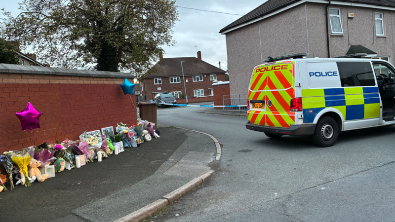 A police van is parked at a cordon. Dozens of tributes, including flowers, cards and toys and pink and blue star-shaped balloons are laid out next to a brick wall.