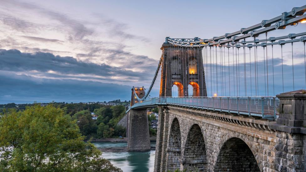 A photo of the Menai Suspension Bridge taken during the evening. The sky is dark and full of clouds. Water can be seen below the bridge. The bridge is lit with warm orange lights. Trees can be seen on each side of the bridge.
