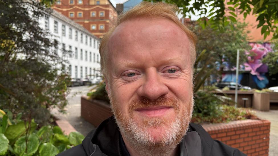 A man with short red hair smiles to the camera. He wears a black coat and stands in front of a large red-brick flowerbed.