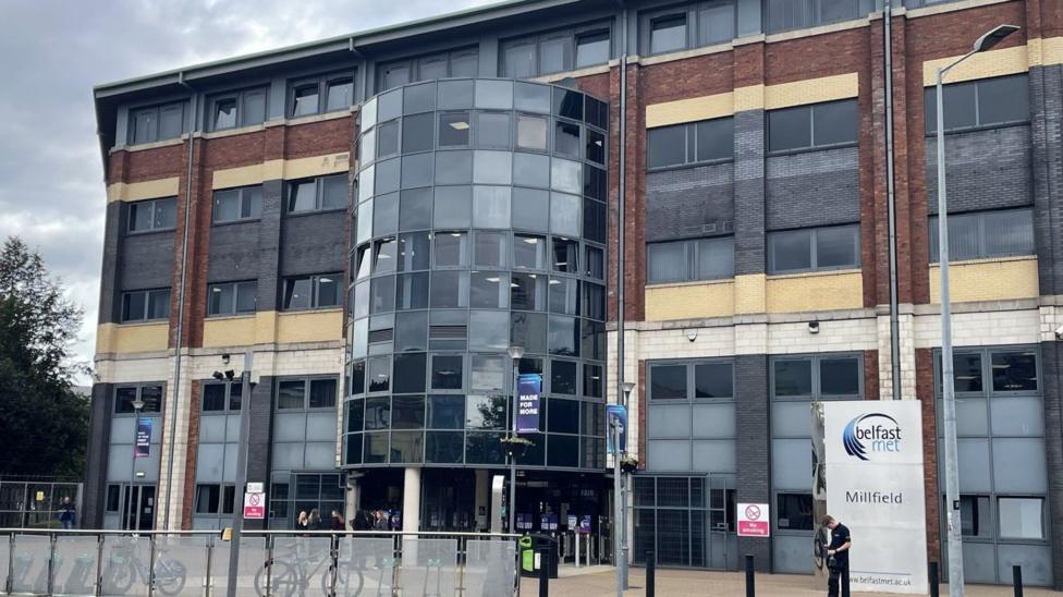 A beige brick building has a black window semi-cylinder entrance. There is a steel grey fence in front of it with bikes leaning on the far side of it. A grey metal sign, to the bottom right of the frame reads "Belfast met"