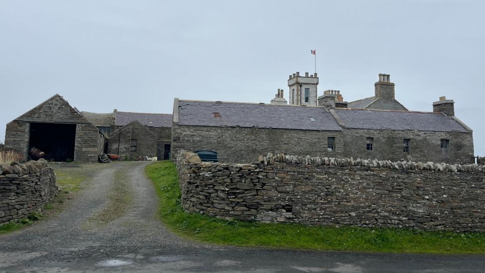 Holland House - a grey brick building on North Ronaldsay. It has several outbuildings and a stone wall. 