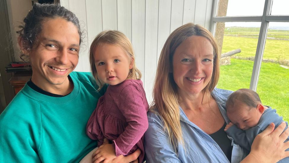 Parents Jorge Suarez and Alice Traill Ford with their son and daughter. Jorge is wearing a green sweatshirt and Alice is wearing a blue overshirt with a black top. Their daughter - a toddler - has on a pink striped dress and baby Duncan is wrapped in a blue blanket. They are standing next to a window and smiling at the camera.