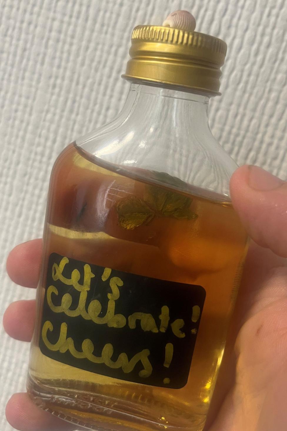 A bottle of whisky with a shell and four-leaf clover glued to it. It has a sticker on it that reads Let's Celebrate! Cheers!