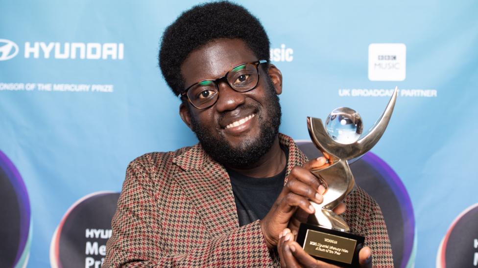 Kiwanuka in a checked suit holding the Mercury Prize trophy and smiling