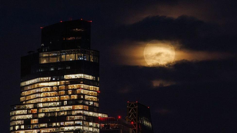 The full Moon is coloured yellowy-orange, with grey clouds shrouding the top and bottom of the Moon. There is a large office building in the foreground with many lights on inside