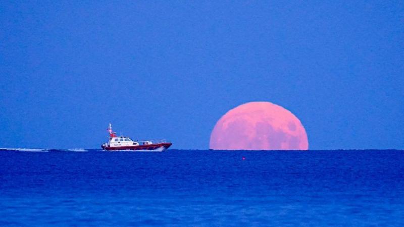 The blue sea goes all the way to the horizon, where half a pink-coloured full Moon is rising into a lighter blue sky. A red and white pilot boat is heading in the direction of the Moon, leaving white surf in its wake